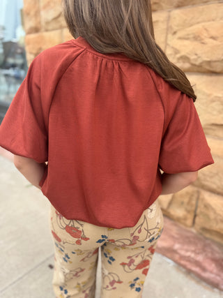 A child viewed from behind wearing a rust-orange short-sleeve top and beige floral pants, standing outdoors against a tan stone wall.