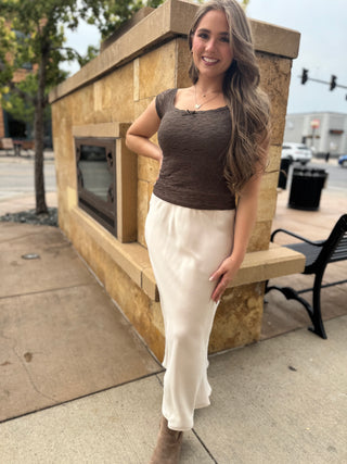 A woman posing outdoors in front of a stone structure, wearing a brown textured scoop-neck top and a cream-colored satin skirt.