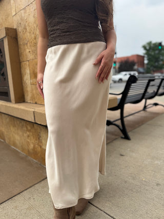 A woman wearing a long cream satin midi skirt with side slits, paired with a dark brown textured top and brown boots, standing outdoors on a city sidewalk.