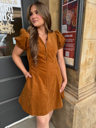 A woman wearing a rust-brown corduroy mini dress with puff sleeves and a front zipper, standing outdoors next to a stone building wall.