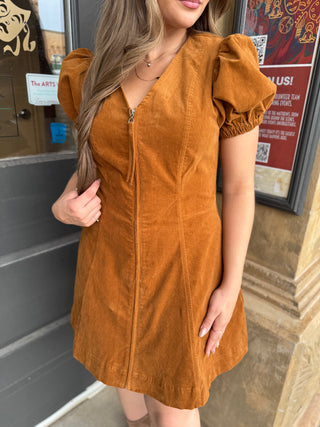 A woman wearing a mustard-brown corduroy mini dress with puff sleeves and a front zipper, standing outdoors in front of a building entrance.