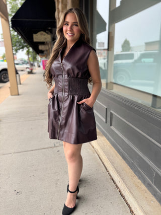 A woman wearing a sleeveless, dark plum faux-leather mini dress with a smocked waist and front zipper, standing on a city sidewalk.