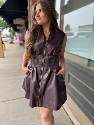 A woman wearing a sleeveless, dark plum faux-leather mini dress with a front zipper, smocked waist, and pockets, standing on a city sidewalk.