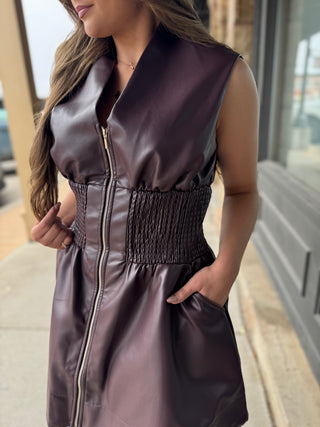 A woman wearing a sleeveless dark plum faux-leather vest dress with a front zipper, smocked waist, and side pockets, photographed outdoors on a sidewalk.