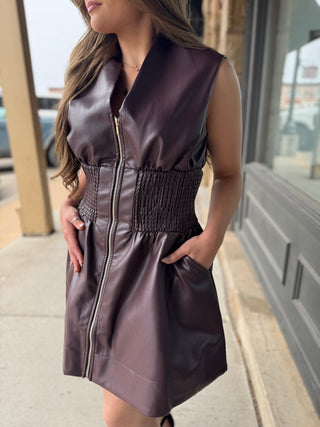 A woman wearing a sleeveless dark plum faux-leather vest dress with a front zipper, smocked waist, and side pockets, standing on a sidewalk.