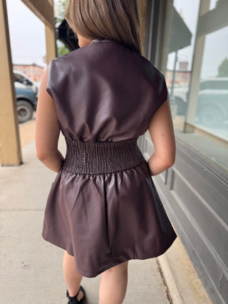 A woman viewed from behind wearing a sleeveless, dark plum-brown faux leather dress with a smocked waistband, standing on a sidewalk next to a storefront.