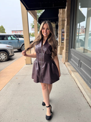 A woman stands on a sidewalk in front of a storefront, wearing a sleeveless dark plum faux-leather mini dress with a front zipper and gathered waist, paired with black heels.
