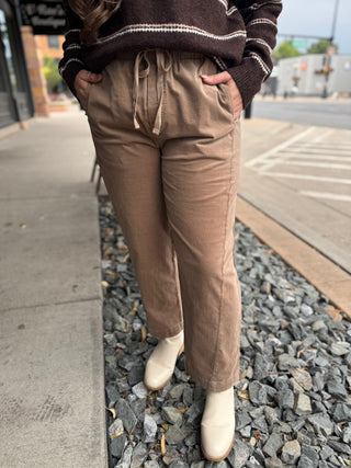 A person standing outdoors on a sidewalk beside a curb, wearing loose brown drawstring pants, a dark brown striped sweater, and off-white ankle boots.
