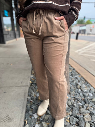 A person wearing tan drawstring wide-leg pants with side pockets, paired with a dark brown sweater and off-white boots, standing outdoors on a sidewalk next to a tree trunk and gravel.
