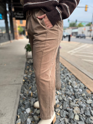 A person wearing tan corduroy drawstring pants, a brown striped sweater, and white boots, standing outdoors on a gravel path next to a sidewalk and street.