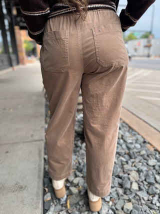 A person viewed from behind wearing loose-fit brown corduroy pants, a dark brown sweater with white trim, and beige shoes, standing outdoors on a sidewalk next to gravel.