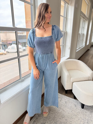 A female model wearing a blue jumpsuit with short puff sleeves, a square neckline, and wide leg design standing indoors.