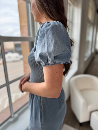 A female model wearing a blue jumpsuit with short puff sleeves, a square neckline, and wide leg design standing indoors.