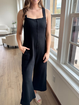 A model wearing a black knit jumpsuit with wide leg and tie detail at the back, standing indoors.