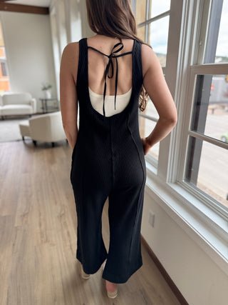 A model wearing a black knit jumpsuit with wide leg and tie detail at the back, standing indoors.