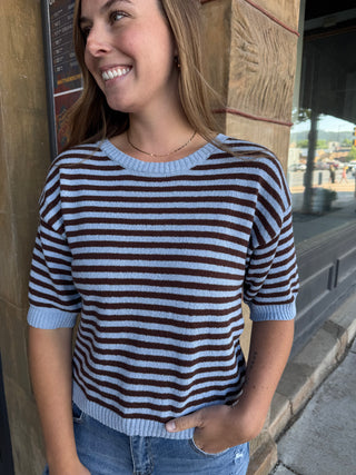 A woman wearing a brown and light blue horizontally striped knit sweater with ribbed trim, standing outdoors next to a wooden pillar.