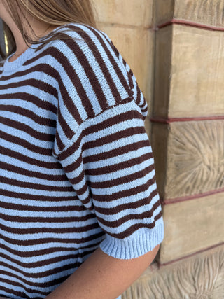 A close-up of a person wearing a short-sleeve knit top with horizontal brown and light blue stripes, standing against a textured beige stone wall.