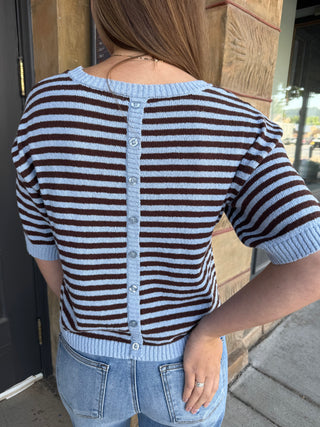 A woman viewed from behind wearing a short-sleeve knit cardigan with horizontal brown and light blue stripes, a light blue button placket, and a ribbed neckline, hem, and cuffs. She is wearing light-wash blue jeans in front of a stone pillar.