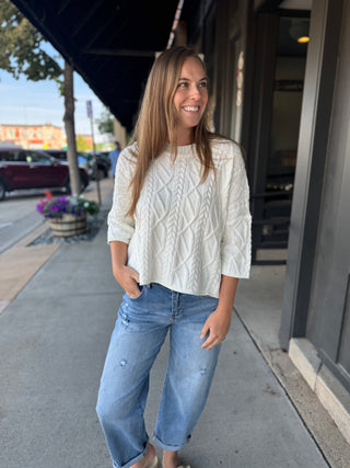 A woman stands on a city sidewalk wearing a cream cable-knit long-sleeve top and light-wash blue jeans.