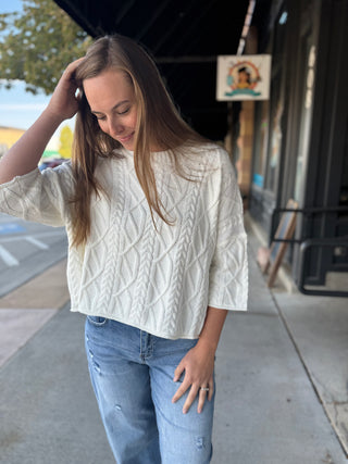 A woman wearing a cream cable-knit short-sleeve top and light-wash blue jeans, standing on a sidewalk in front of a storefront.