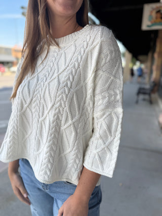 A woman wearing a cream cable-knit cropped sweater with three-quarter sleeves, paired with blue jeans, standing outdoors on a sidewalk.