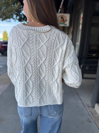 A woman viewed from behind wearing a cream cable-knit sweater and light blue jeans, standing on a sidewalk in front of a storefront.