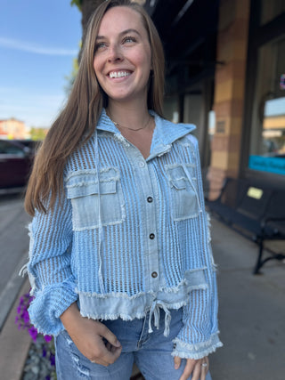 A woman smiling outdoors while wearing a light blue distressed denim jacket with a textured open-knit pattern, button front, and frayed edges.