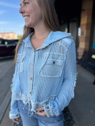 A woman wearing a light blue distressed mesh hooded button-up shirt with frayed details and denim-style pockets, photographed outdoors on a sidewalk.
