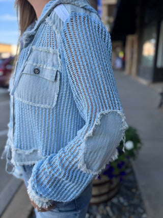 A person wearing a light blue open-knit cropped jacket with distressed frayed edges and light blue denim patches on the sleeves, photographed outdoors on a sidewalk.
