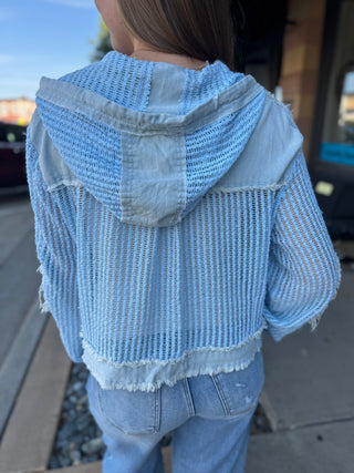 A person viewed from behind wearing a light blue open-knit hooded jacket with frayed edges and light-wash jeans, standing outdoors on a sidewalk.