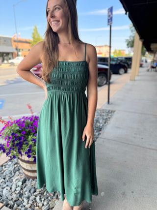 A woman wearing a sage green midi dress with a smocked bodice and thin spaghetti straps, standing on a sidewalk in front of a storefront.