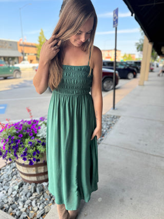 A woman wearing a sage green smocked midi dress stands on a sidewalk in front of a storefront. The dress has thin straps, a smocked bodice, and a flowing skirt with pockets.