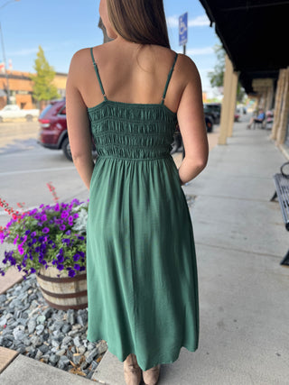 A woman viewed from behind wearing a sage green midi dress with thin spaghetti straps and a smocked bodice, standing on a sidewalk in front of a storefront.