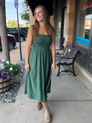 A woman wearing a sage green smocked strapless midi dress and beige ankle boots stands on a sidewalk in front of a storefront.