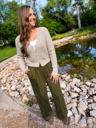 A woman stands outdoors by a rocky pond wearing a light beige knit cardigan over a white top and olive green drawstring pants.