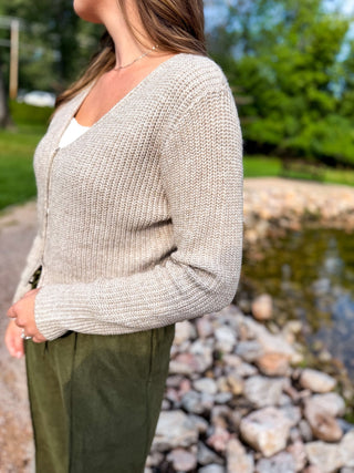 A woman viewed from behind wearing a light beige textured knit sweater and olive green pants, standing outdoors near a rocky body of water.