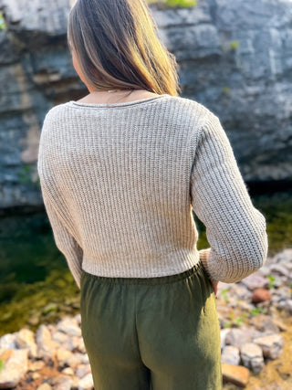 A woman viewed from behind wearing a light beige textured knit sweater and olive green pants, standing outdoors near a rocky body of water.
