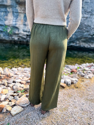A person standing outdoors by a rocky shoreline wearing loose olive green wide-leg pants and a cream knit sweater, photographed from the back.