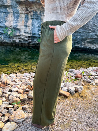 A person standing outdoors by a rocky shoreline wearing olive green wide-leg pants and a beige ribbed knit sweater, with water and a cliff in the background.