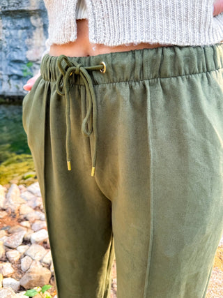 Olive green drawstring sweatpants worn by a model outdoors near a stone and water background.