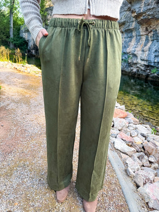A person wearing olive green wide-leg drawstring pants, standing outdoors on a gravel path next to a rocky stream with trees and cliffs in the background.