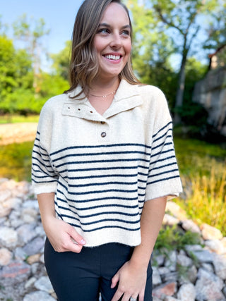A woman wearing a cream knit polo sweater with navy horizontal stripes and a button collar, standing outdoors near a rocky pond with trees in the background.