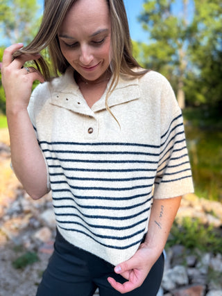 A woman wearing a cream knit polo sweater with navy horizontal stripes, standing outdoors near a pond with trees in the background.