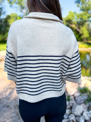 A person viewed from behind wearing a cream knit cardigan with navy horizontal stripes across the lower body and sleeves, photographed outdoors near trees and rocks.