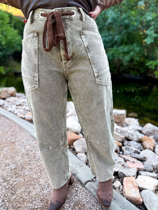 A person wearing light beige corduroy pants with a brown drawstring waist, standing outdoors on a stone path near water and greenery.
