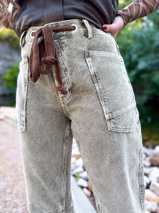 Close-up of a person wearing light beige corduroy pants with a brown knotted drawstring at the waist, standing outdoors near rocks and greenery.