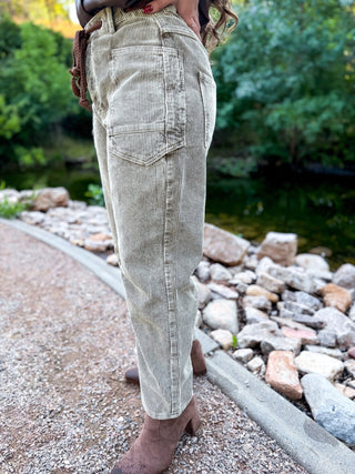 A person wearing light beige corduroy pants and brown boots, standing outdoors on a gravel path next to a rocky stream with green trees in the background.