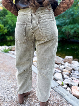 A person viewed from behind wearing light beige corduroy pants, standing outdoors on a gravel path next to a stone-lined water feature with green foliage in the background.