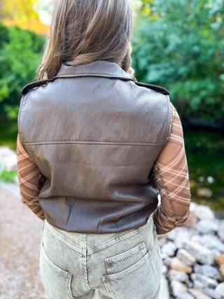 A person viewed from behind wearing a dark brown faux-leather vest over a brown and beige plaid long-sleeve shirt and light gray corduroy pants, standing outdoors near a rocky stream with green foliage in the background.
