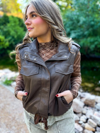 A woman wearing a brown faux-leather vest over a brown plaid shirt, standing outdoors near a stone path and greenery.